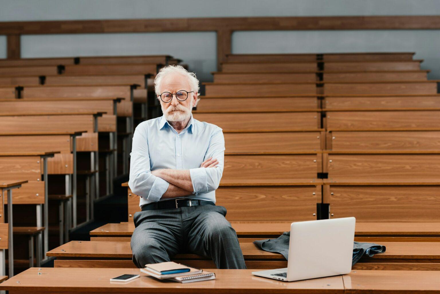 grey hair professor sitting in empty lecture room with crossed arms