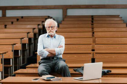 grey hair professor sitting in empty lecture room with crossed arms