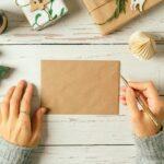 Woman hands in sweater writing on blank envelop greeting card . Flat lay of white wooden background