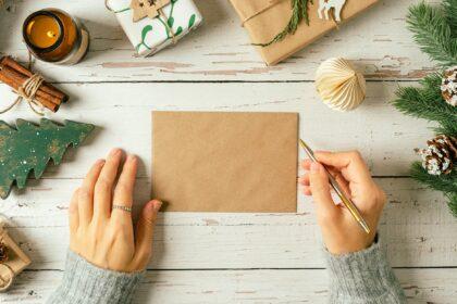 Woman hands in sweater writing on blank envelop greeting card . Flat lay of white wooden background