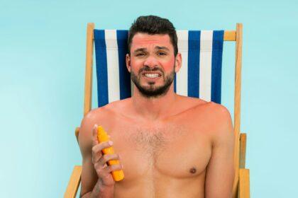 Worried shirtless man with sunburn holding sunscreen isolated on blue