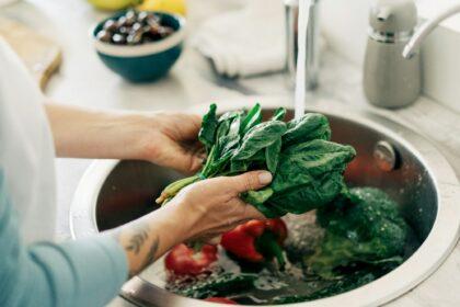 An unrecognizable woman washes greens and vegetables under tap water in the kitchen sink.