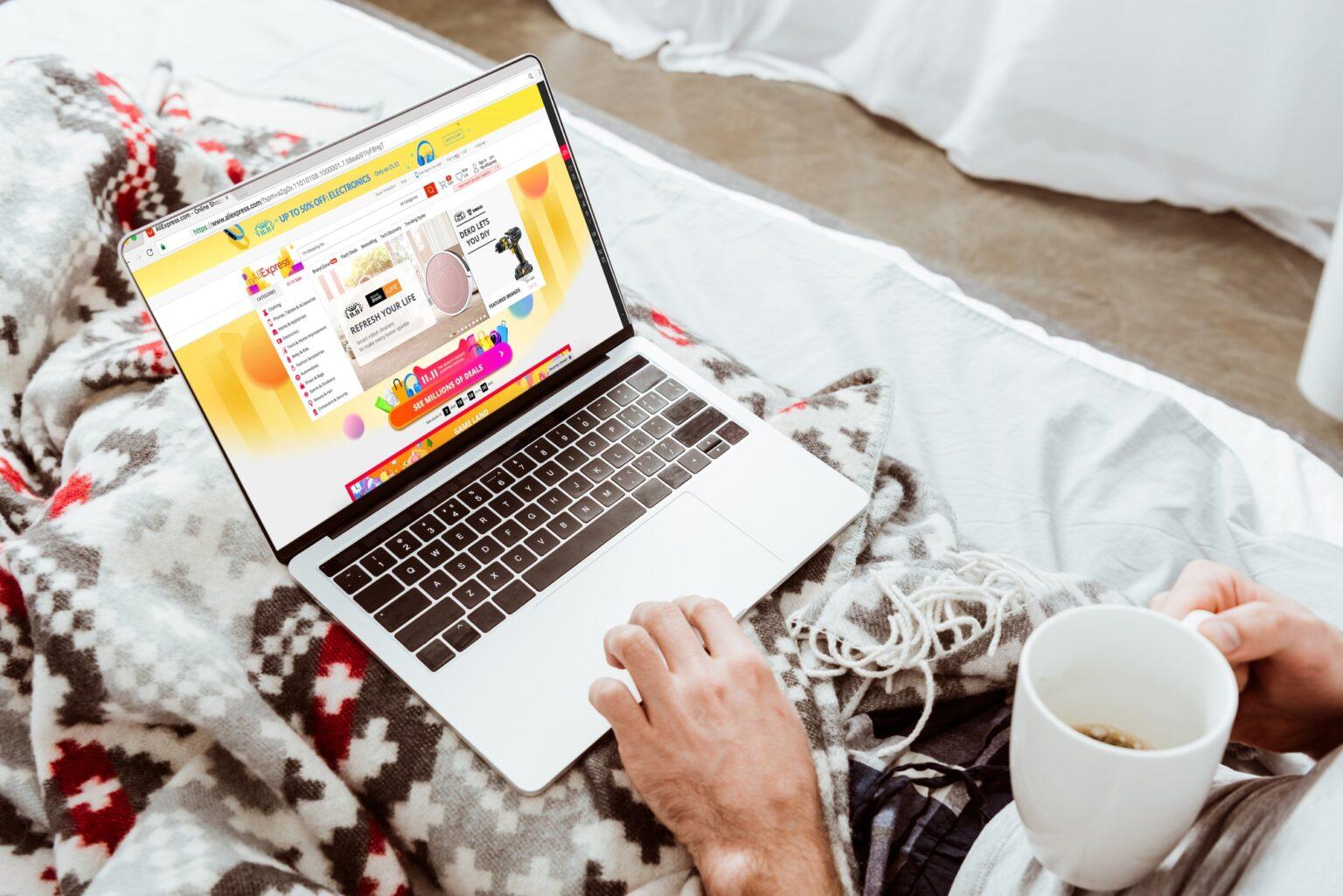 cropped image of man holding coffee cup and using laptop with aliexpress on screen in bed at home