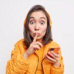 Studio shot of surprised woman gossips about something makes silence gesture tells secret holds mode