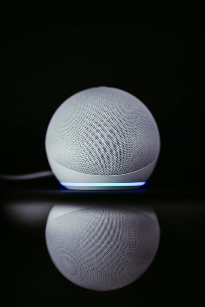 A modern smart speaker Echo Dot with a sleek design and glowing base, reflected on a dark surface.
