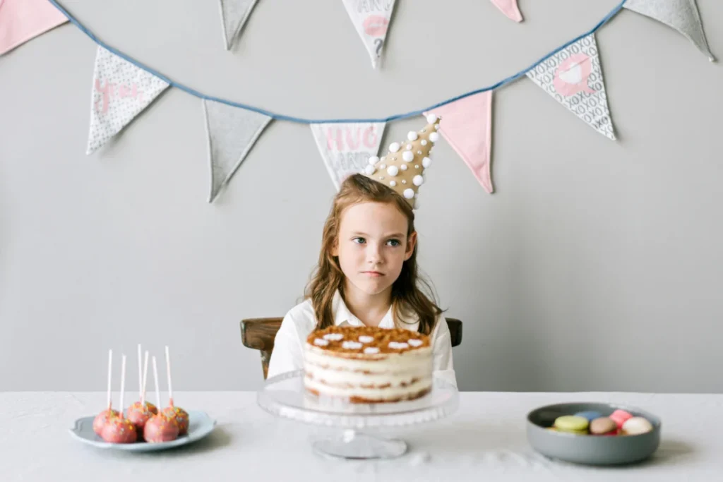 A young girl wearing a party hat sits at a table with a birthday cake and treats.