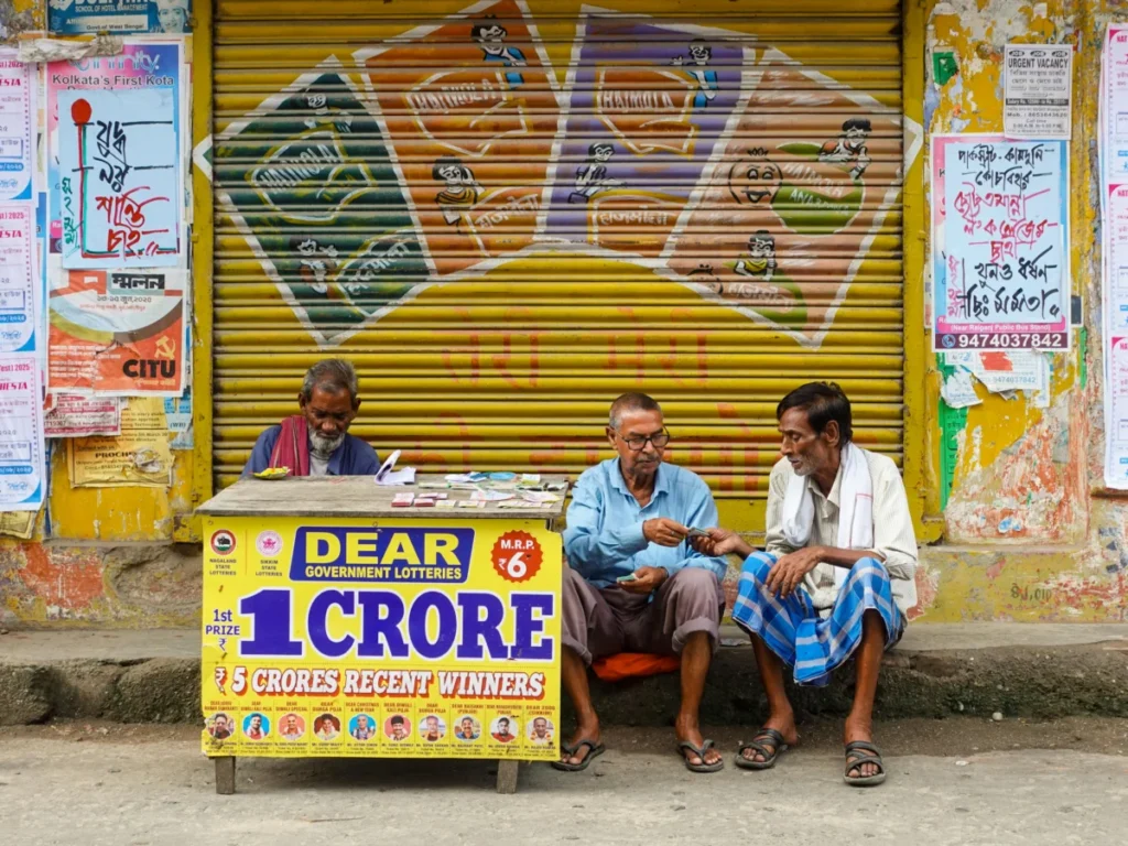 Three men sit at a lottery booth outside in Raiganj, India. Colorful posters and signs add vibrancy.